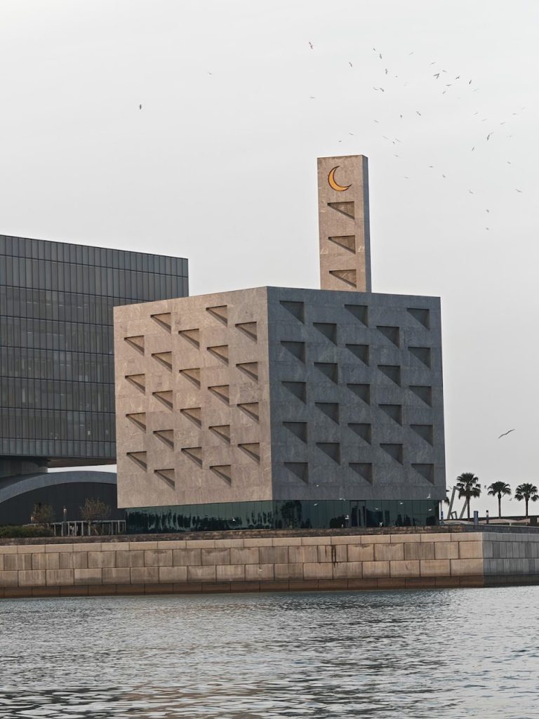 Waterfront mosque volume with geometric openings, pale sky, and water in Bahrain.