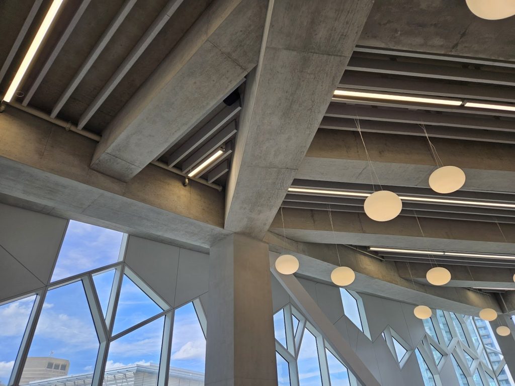 Concrete beams, suspended lights, and angled glazing forming an interior ceiling field.